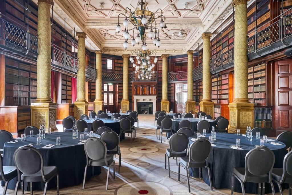 Elegant dining setup inside a grand library-style room with ornate columns, tall bookshelves filled with colorful books, and decorative chandeliers hanging from a detailed ceiling. Round tables are arranged neatly across the patterned carpet