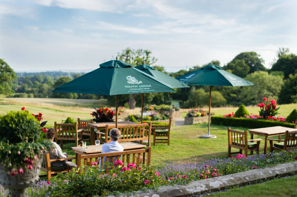 Wooden tables and chairs in a neat garden, with a fantastic view. Two people are sitting at a table with drinking glasses.